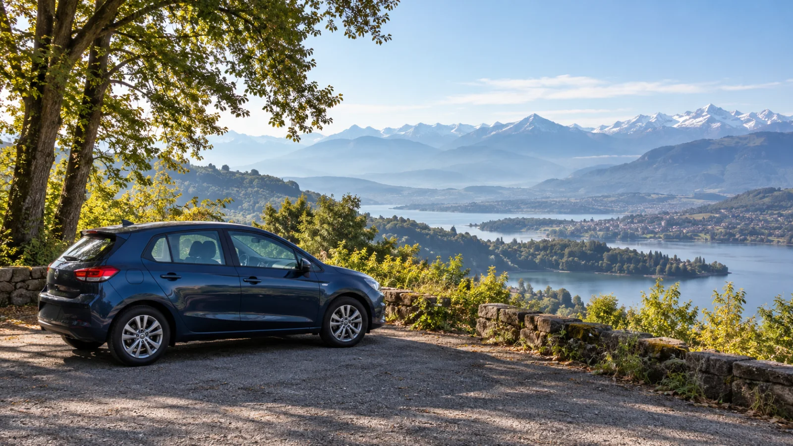Blue compact car at a wooded viewpoint above a lake and distant mountains